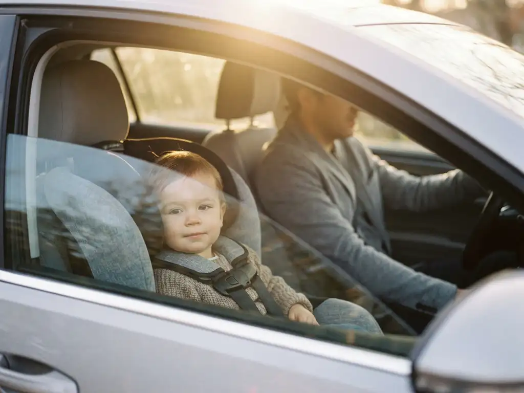 Young child buckled in front passenger seat with parent driving, warm afternoon sunlight through windshield, calm family car ride.