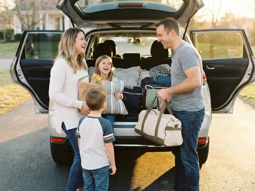 Fröhliche Familie belädt einen silbernen SUV mit Snacks, Kissen und Reisetasche in sonniger Einfahrt bei goldenem Abendlicht.
