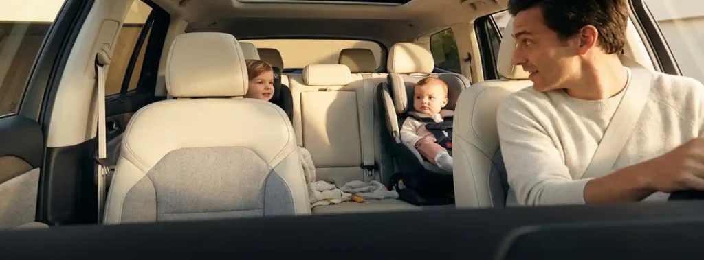 Young family inside a modern car, children secured in rear car seats, parent glancing back warmly, golden sunlight through windows.