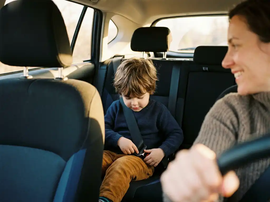 Young child buckling their own seatbelt in the backseat while a parent smiles encouragingly from the front seat.